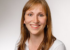 Porträt einer lächelnden Frau mit langen, braunen Haaren, die einen weißen Hemdkragen trägt. Der Hintergrund ist hell und neutral. Ihr Gesichtsausdruck ist freundlich und einladend.