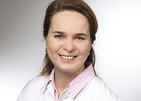 Porträt einer lächelnden Frau mit langen, braunen Haaren, die ein weißes Hemd mit rosa Kragen trägt. Der Hintergrund ist hell und neutral gehalten. Die Frau strahlt Freundlichkeit und Professionalität aus.