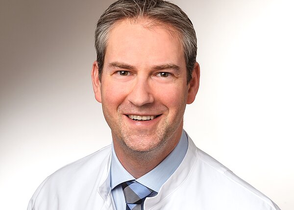 Das Bild zeigt einen lächelnden Mann in einem weißen Arztkittel. Er hat kurzes, graues Haar und trägt ein hellblaues Hemd mit einer karierten Krawatte. Der Hintergrund ist hell und neutral.