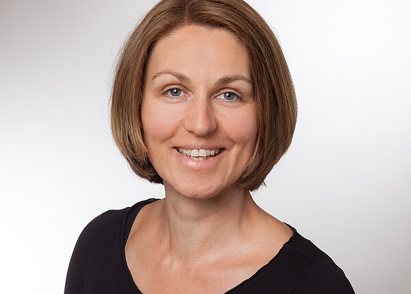 Porträt einer lächelnden Frau mit kurzen, braunen Haaren. Sie trägt ein schwarzes Oberteil und steht vor einem hellen Hintergrund. Die Frau hat ein freundliches und offenes Ausdruck, das eine positive Atmosphäre vermittelt.